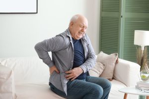 A man is sitting on the couch suffering from back pain and will visit Schererville Chiropractor to alleviate the pain.